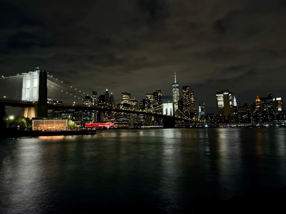 Beleuchtete Manhattan-Skyline und Brooklyn Bridge vom Ufer bei Nacht