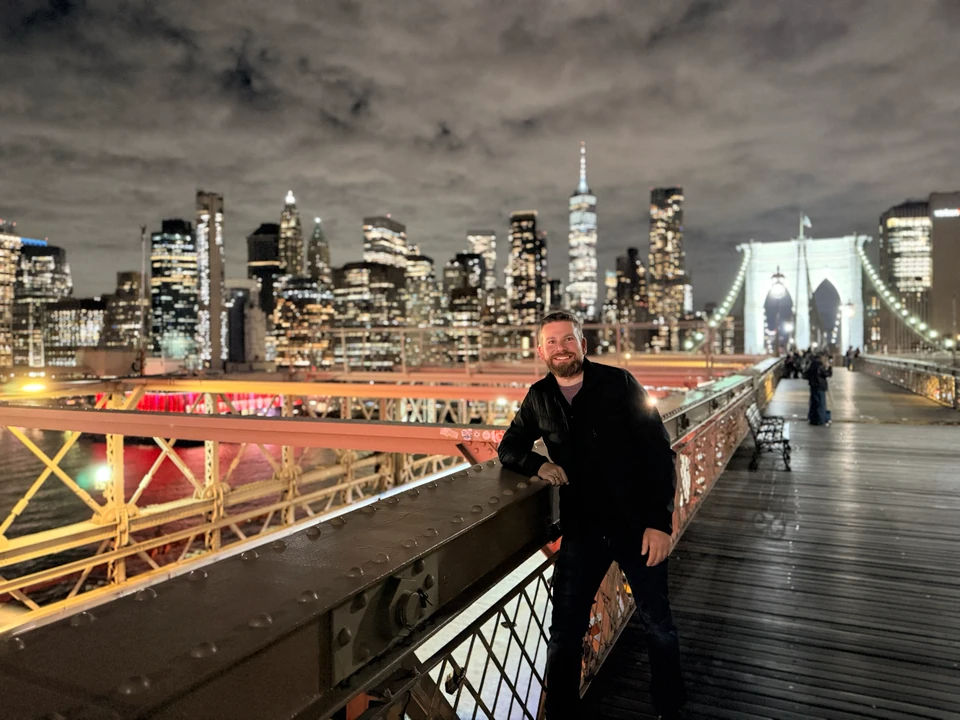 Tobias auf dem Fußweg der Brooklyn Bridge vor der nächtlichen Manhattan-Skyline
